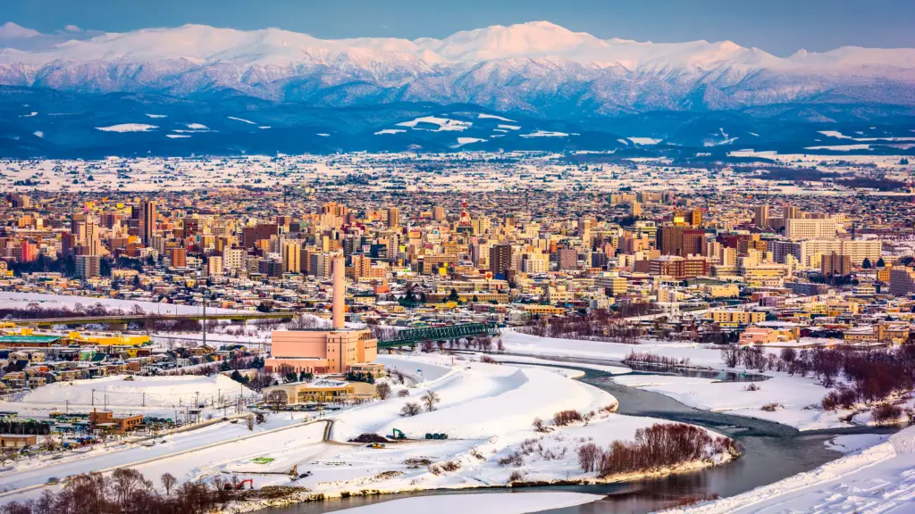 Scenic winter view of a city in Hokkaido, Japan, with snow-covered landscapes, a winding river, and distant mountains in the background.