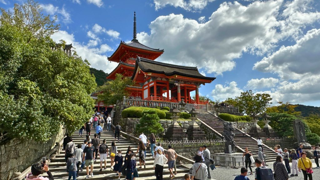 Tourists walking up the stone steps leading to Kiyomizu-dera Temple in Kyoto, Japan. The vibrant red pagoda and temple gate stand against a backdrop of lush greenery and a bright blue sky, showcasing the beauty of this iconic UNESCO World Heritage Site.