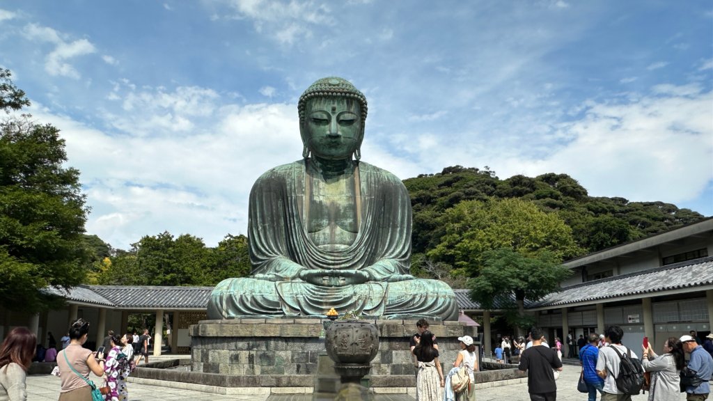 The Great Buddha of Kamakura (Daibutsu) at Kotoku-in Temple, a massive bronze statue standing 11.4 meters tall, surrounded by visitors against a backdrop of lush greenery and blue skies.