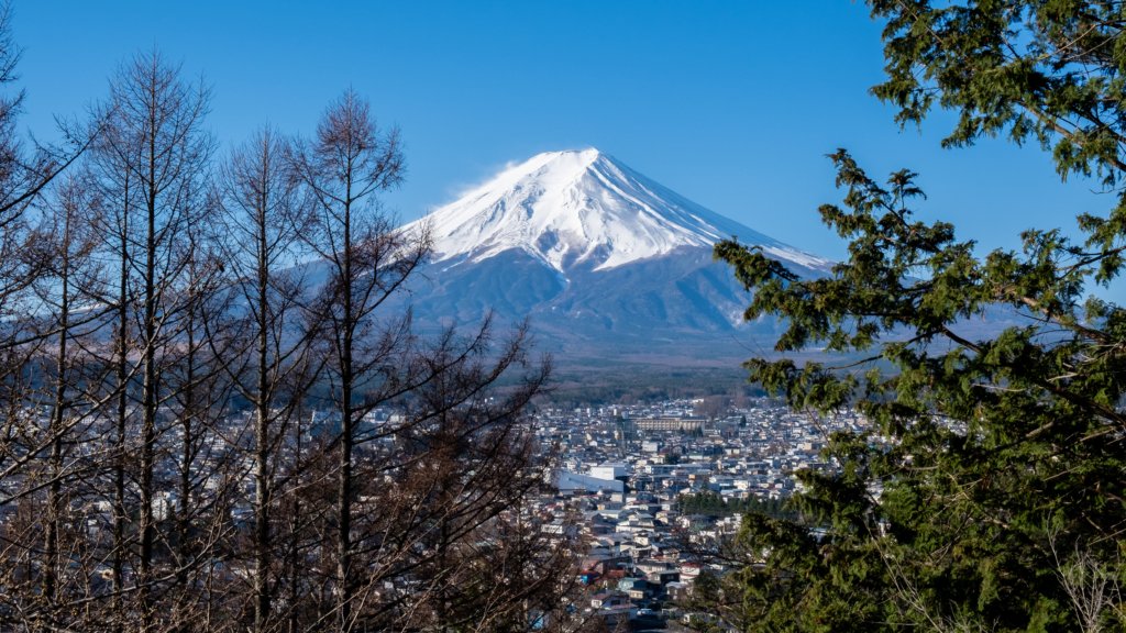 Stunning view of Mount Fuji with its snow-capped peak, framed by trees and overlooking a cityscape at its base in Japan.