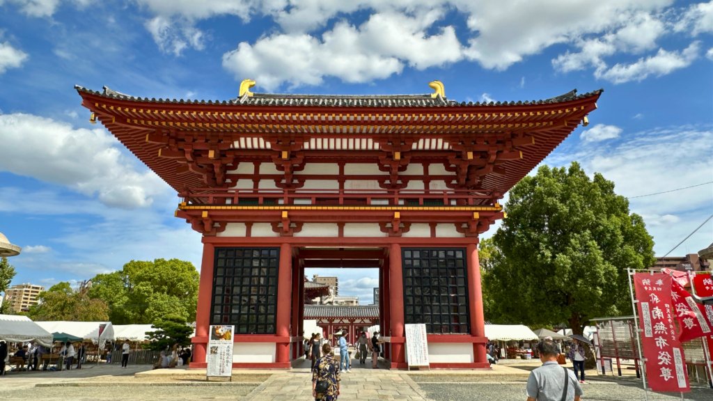 The grand entrance gate of Shitenno-ji Temple in Osaka, Japan, showcasing its traditional red and white architecture under a bright blue sky with scattered clouds.