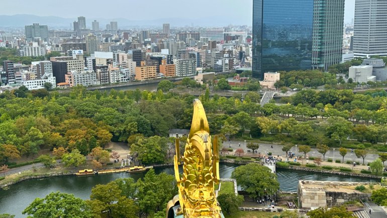 View from the top of Osaka Castle overlooking the city skyline, lush greenery, and the castle moat, with a golden shachihoko (ornamental fish) in the foreground.