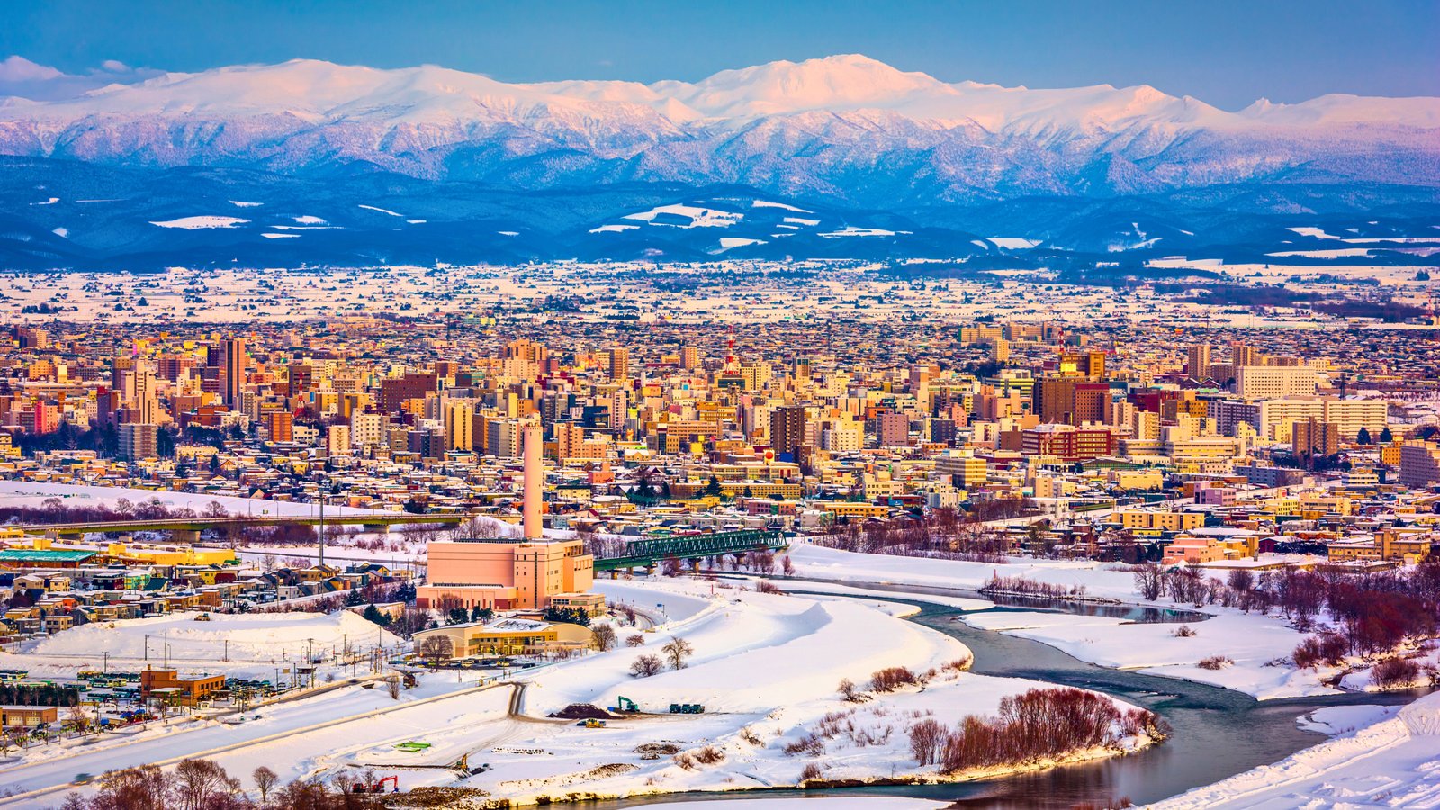 A breathtaking winter view of Asahikawa, Hokkaido, Japan, with snow-covered landscapes, a winding river, and a backdrop of majestic mountains.