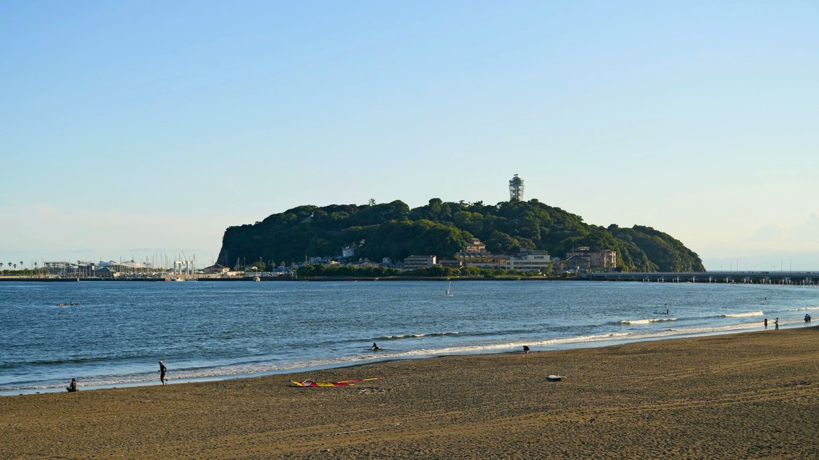 Enoshima Island, Japan A scenic view of Enoshima Island, Japan, from the sandy beach, with the Enoshima Sea Candle observation tower rising above lush greenery and the connecting bridge in the background.