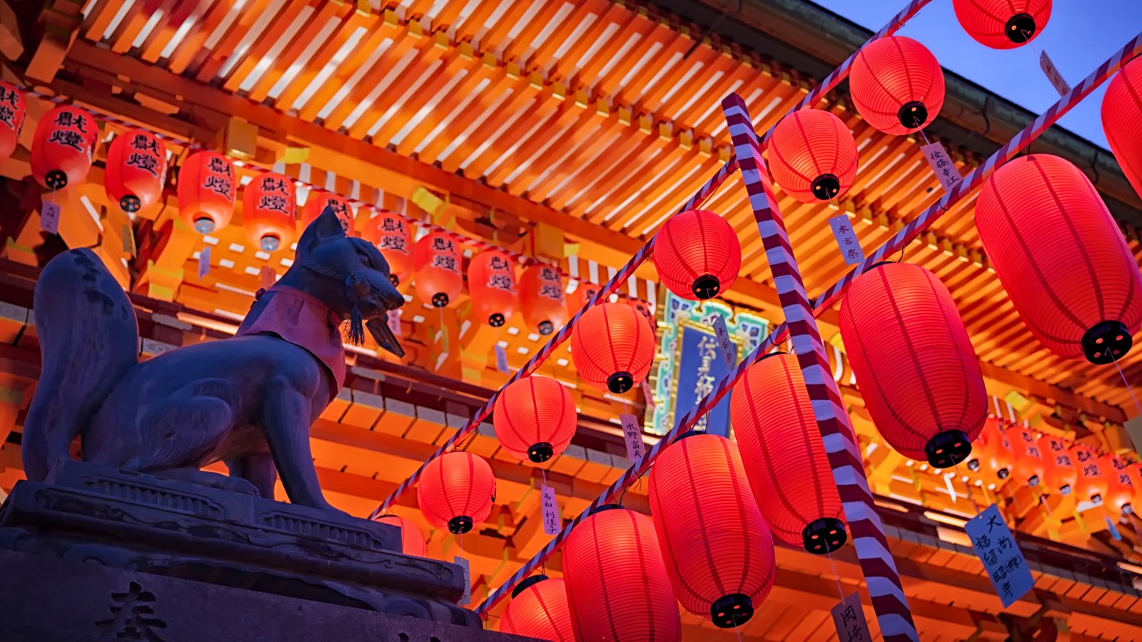 Illuminated red lanterns and a fox statue at Fushimi Inari Taisha Shrine in Kyoto, Japan, during the Motomiya Festival, creating a vibrant nighttime scene.