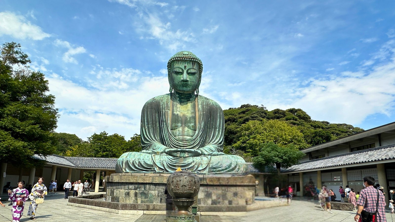 The Great Buddha of Kamakura, a massive bronze statue standing 13.35 meters tall, surrounded by visitors and lush greenery under a bright blue sky.